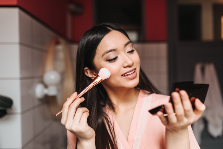 young-brunette-woman-applies-makeup-with-brush-looks-into-small-mirror